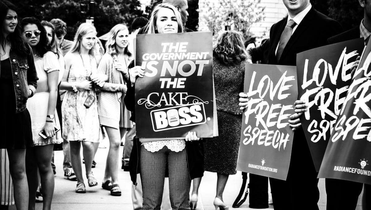 A black and white photograph of a crowd of protestors. A young woman holds a sign that says, "The government is not the cake boss." Other signs read, "Love free speech."