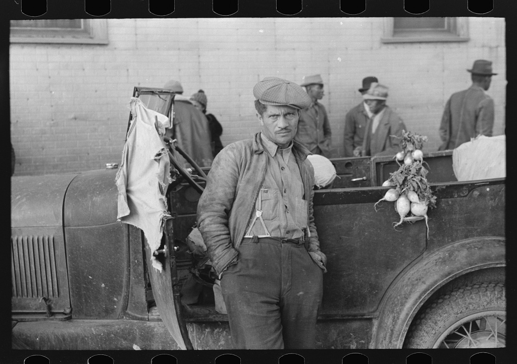 An unnamed fair skinned man, working as a vegetable vendor, dressed in a collared shirt, suspenders, dark pants, and an early 20th century style flat cap, leans against an old automobile next to a bunch of turnips. In the background, out of focus, there are African American men, also in hats, walking by.