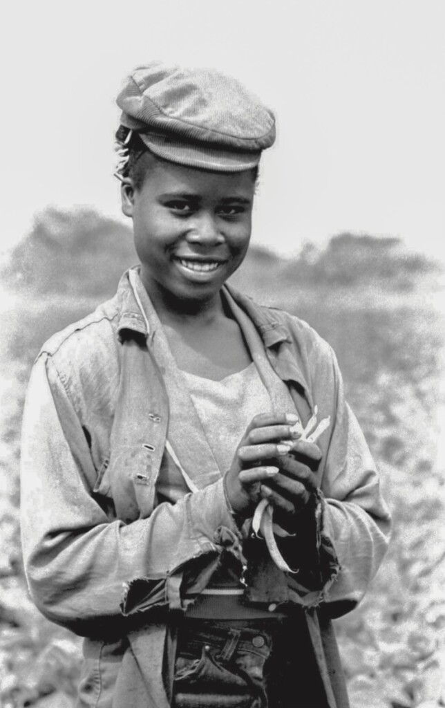 A young African American woman stands in a field wearing a corduroy hat, a light jacket with small tears at the wrist, and faded jeans. She smiles at the camera while holding a handful of freshly picked snap beans.