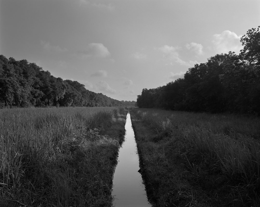 A vast open field lines with trees and a single pale strip of water cutting through the center towards the sky.