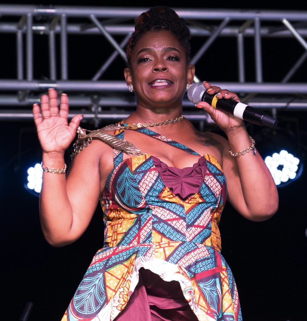 Portrait of Asali DeVan Ecclesiastes in colorful dress, holding microphone.