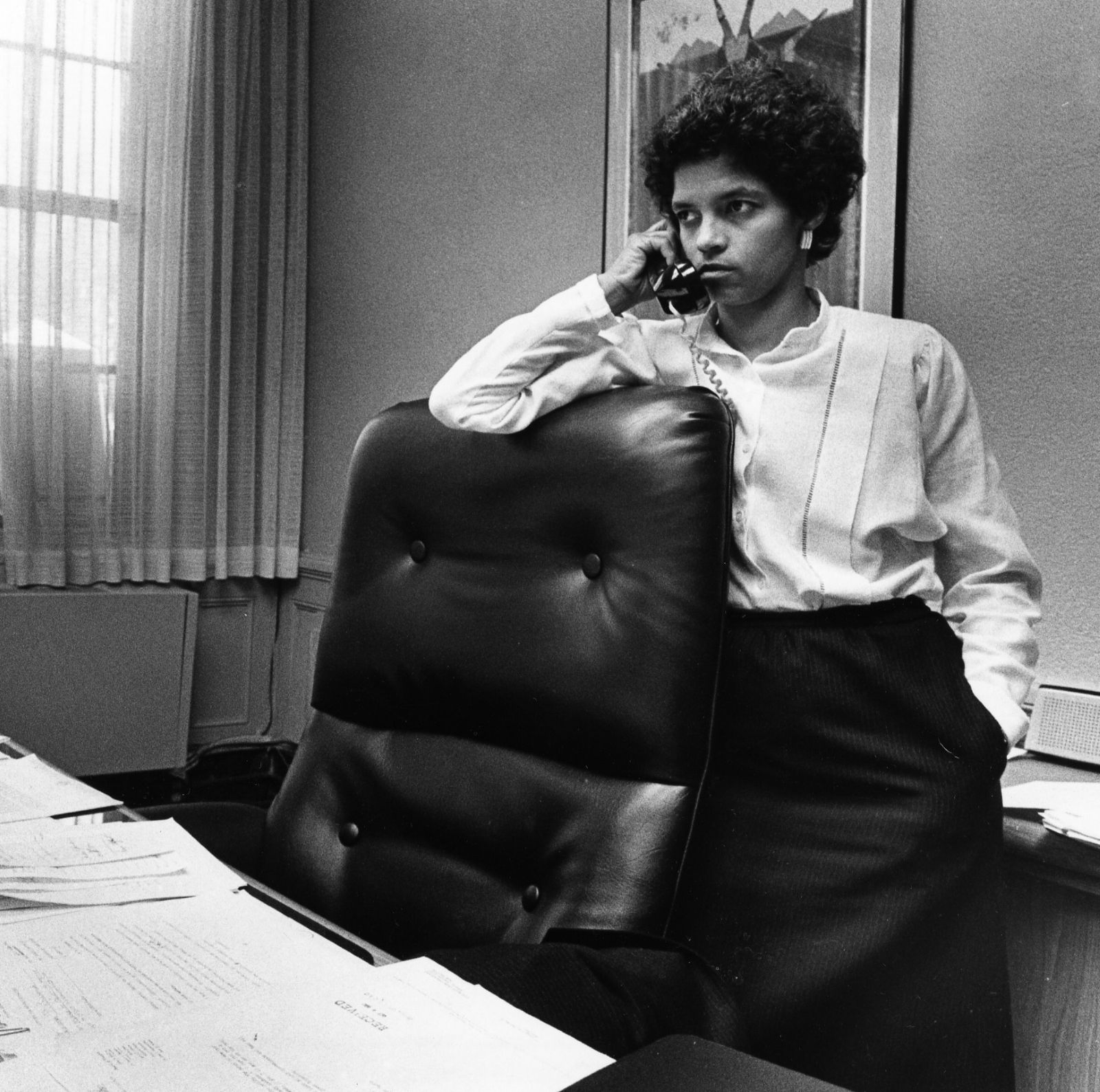 Shirley Franklin stands behind an office chair with a telephone to her ear. Papers are spread across the desk in front of her.
