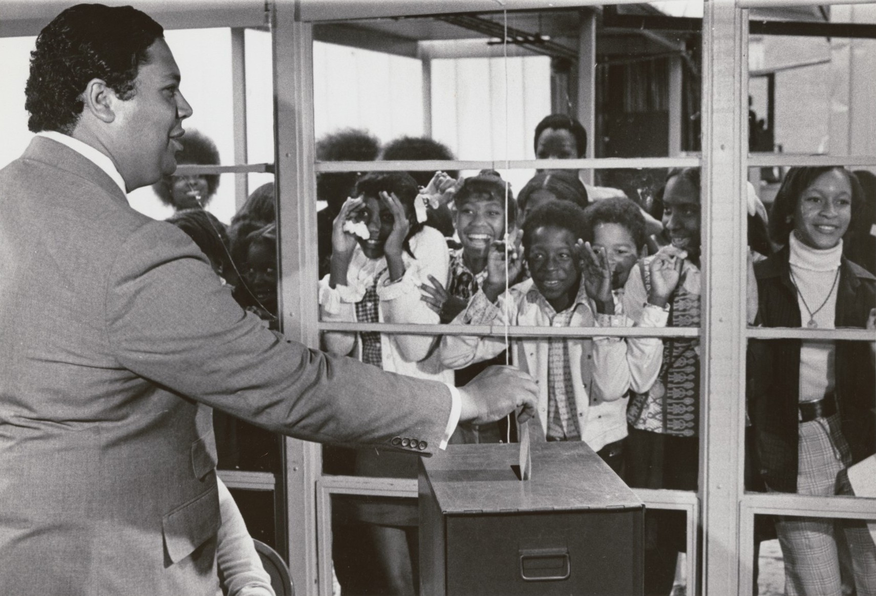 Maynard Jackson casts his vote as a group of smiling Black children watch through a window.